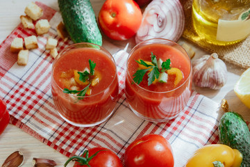 Tomato soup gazpacho in glass with herbs and ingredients on the kitchen. A beautiful summer soup spilled in glasses surrounded by juicy vegetables.