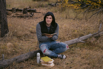 A young handsome man in casual clothes is sitting on a log and reading a book in the autumn forest.