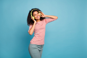 Photo of cheerful cute curly nice toothy attractive gorgeous black skinned woman holding her headphones wearing trousers pants listening to music isolated blue vivid color background