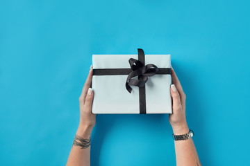 Hands of woman with tattoes and watch holding white paper gift box tied with black ribbon and bow on blue background. Close-up, copy space, top view.