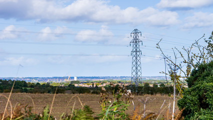 Image of a cultivated field with an electric pylon in the background with blurred foreground elements framing the shot. Taken on a warm autumn day with a blue sky and white clouds.