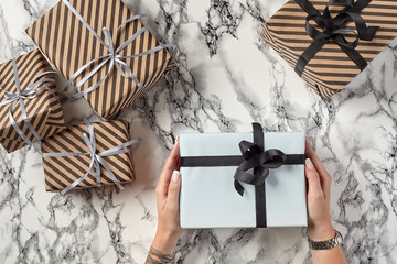 Hands of a woman are holding white paper gift box tied with black ribbon and bow against a marble background. Close-up, copy space, top view.