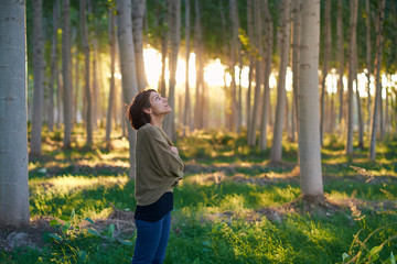 Latin brunette woman walks among the trees of a forest at sunset. The sun's rays enter through the vegetation. She wears a brown sweater, a black T-shirt and a jeans.