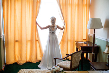 The blonde bride in a boudoir dress opens the curtains in the hotel room and looks out the window while preparing for the wedding and waiting for the groom. Inspiration, love, feelings, happiness.