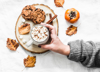 Woman's hand in a warm sweater holds cup of hot chocolate. Cookies with chocolate chips and mug of hot chocolate on a light background, top view. Cozy autumn winter still life