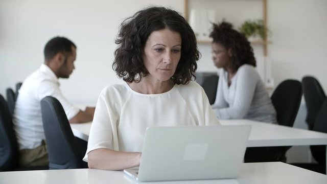 Focused mature woman working with laptop in office. Thoughtful middle aged lady using modern device. Technology concept