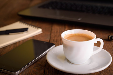 Cup of coffee on a laptop keyboard. Work place modern. Laptop (notebook) with cup of coffee and notepad with pen on old wooden table.