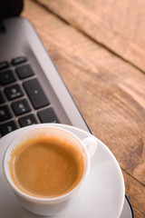 Cup of coffee on a laptop keyboard. Work place modern. Laptop (notebook) with cup of coffee and notepad with pen on old wooden table.