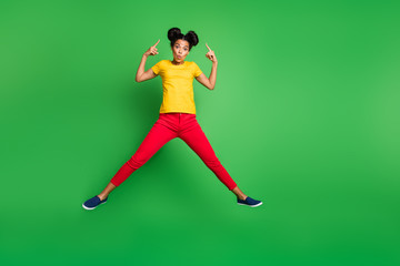 Fototapeta na wymiar Full size photo of pretty dark skin lady jumping high indicating fingers on new stylish hairstyle wear casual yellow t-shirt red pants isolated green background