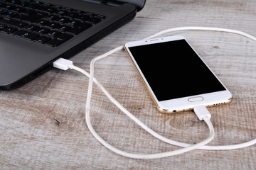 A gray laptop and a white telephone(smartphone) connected by a cable for data transmission on a wooden table. Data transfer. Information exchange