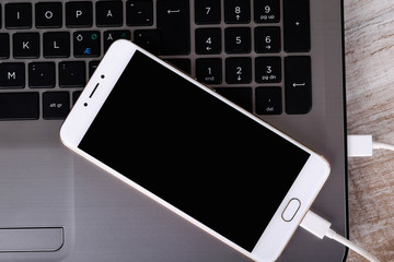 A gray laptop and a white telephone(smartphone) connected by a cable for data transmission on a wooden table. Data transfer. Information exchange