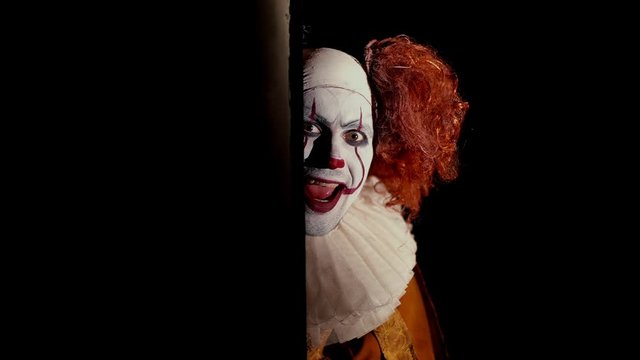 Crazy clown with a wide, frightening smile slowly appears from a dark corner. Shooting in black room, close-up face