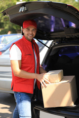 Young courier holding parcels near delivery car outdoors
