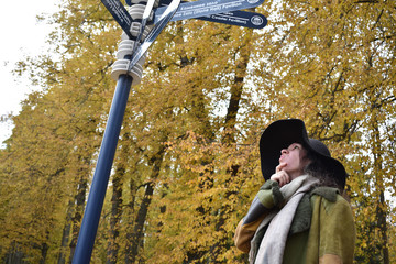 young beautiful pretty lady woman posing in a long green and brown dress gown overcoat walks down a lonely alley in the garden and looks at the information sign with emotions