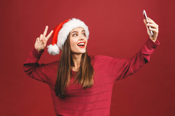Christmas portrait of beautiful girl. Teenager wearing santa claus hat. Girl smiling and making selfie on mobile phone isolated against red background.