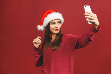 Christmas portrait of beautiful girl. Teenager wearing santa claus hat. Girl smiling and making selfie on mobile phone isolated against red background.