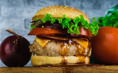 A huge juicy hamburger is on a chopping board with vegetables on a gray brick background.