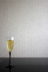 Alone wineglass with white wine on black glass table. Gray blurred wall behind. Background with yellow liquid in goblet and place for text.