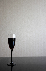 Alone wineglass with red wine on black glass table. Gray blurred wall behind. Background with dark liquid in goblet and place for text.