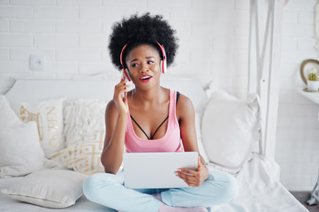 Young african american woman sitting in bed while working on laptop, speaking at mobile phone and listen music on earphones.