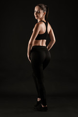 Obraz premium Brunette woman in black leggings, top and sneakers is posing against a black background. Fitness, gym, healthy lifestyle concept. Full length.