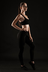 Fototapeta na wymiar Brunette woman in black leggings, top and sneakers is posing against a black background. Fitness, gym, healthy lifestyle concept. Full length.