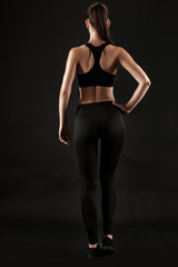 Fototapeta premium Brunette woman in black leggings, top and sneakers is posing against a black background. Fitness, gym, healthy lifestyle concept. Full length.