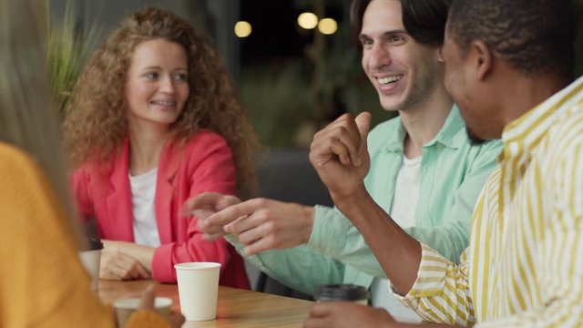 Happy young man with cup of fresh coffee gesturing and speaking with multiracial friends while resting in cafe