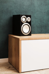 Single modern black audio speaker in interior on wooden desk near the wall.