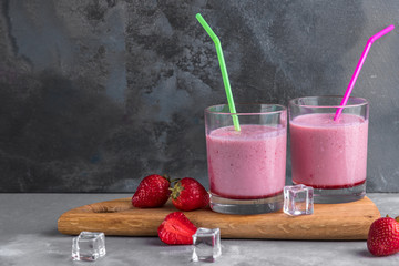 two glasses of strawberry cocktail with straws on a wooden board and gray background with ice cubes, refreshment drinks for summer, copy space fot text