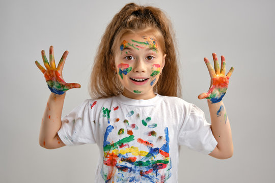 Little girl in white t-shirt is posing standing isolated on white and gesticulating with her painted in different colors palms. Art studio. Close-up.