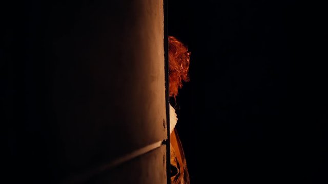 Scary clown with a crazy smile on his face suddenly appears around the corner in darkroom. Terrible clown with a colorful makeup in a festive costume. Shooting in slow motion