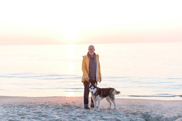 Elderly man with grey hair and beard in a yellow raincoat with husky dog on leash walking on the beach
