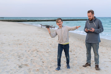 Bearded man and handsome brunette boy with stylish haircut which holds drone like airplane. Father and son with drone on the beach 