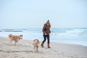 Young woman with long red hair in hat and jacket walking husky dog on seaside