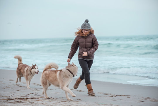 Young woman with long red hair in hat and jacket walking husky dog on seaside