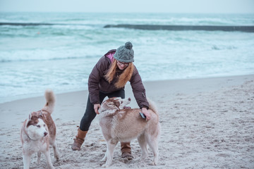 Young woman with long red hair in hat and jacket walking husky dog on seaside