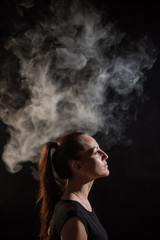 The brunette smokes an electronic cigarette on a black background, releases thick smoke from her mouth. Portrait of a woman smoking a vape, hovering.