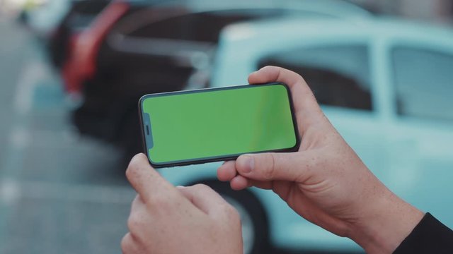 Close-up of male hands browsing application on horizontal mock-up smartphone greenscreen outdoors on background with parked cars.