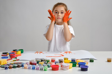 Little girl in white t-shirt sitting at table with whatman and colorful paints on it, painting her cheeks. Isolated on white. Medium close-up.