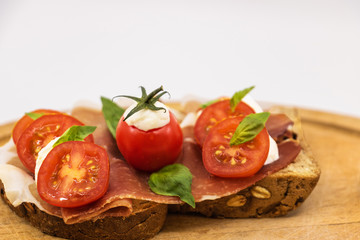 Sandwich italien sur une tranche de pain multi céréales avec une tranche de jambon rouge des tomates cerises des billes de mozzarella et du basilic frais sur une assiette en bois -isolé fond blanc