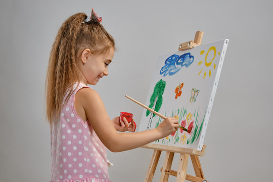 Little girl artist in a pink dress is standing behind easel and painting with brush on canvas, isolated on white studio background. Medium close-up.