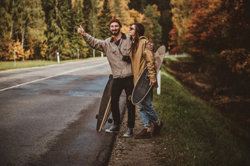 Fototapeta premium Couple of happy smiling students with longboards are trying to catch up car in the autumn park.