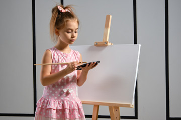 Little girl artist in a pink dress is standing behind easel and painting with brush on canvas at art studio with white walls. Medium close-up shot.