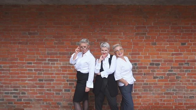 Three adult caucasian blondes businesswoman in office suits pose near brown brick wall. Two of them arms crossed, one of them keep hand in pocket