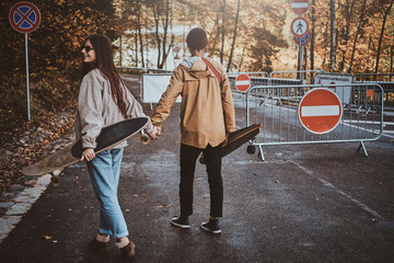 Couple of young romantic hipsters are enjoying their walk at autumn park while holding their longboards.