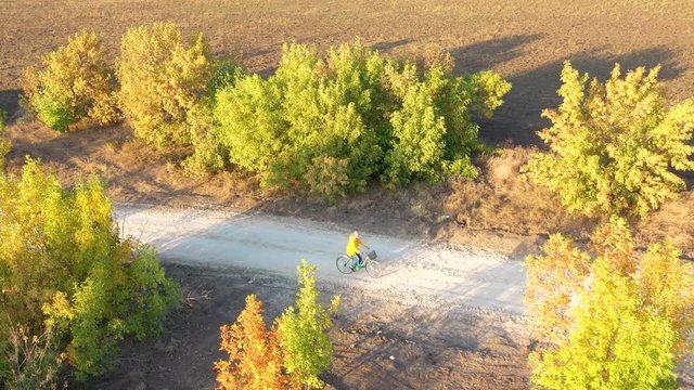 Aerial view of man in yellow raincoat is riding along rural road and scorched earth in autumn forest at sunset. Drone video of contryside landscape and rider on cycle