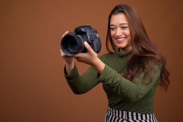 Young beautiful Indian woman with camera against brown backgroun