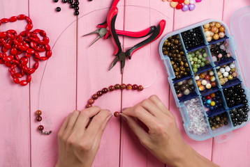 Jewelry making. Making bracelet of colorful beads. Female hands with a tool on a pink wooden table. - obrazy, fototapety, plakaty