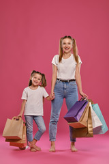 Mom and daughter with ponytails, dressed in white t-shirts and blue jeans are posing against pink background with packages in hands. Full length.